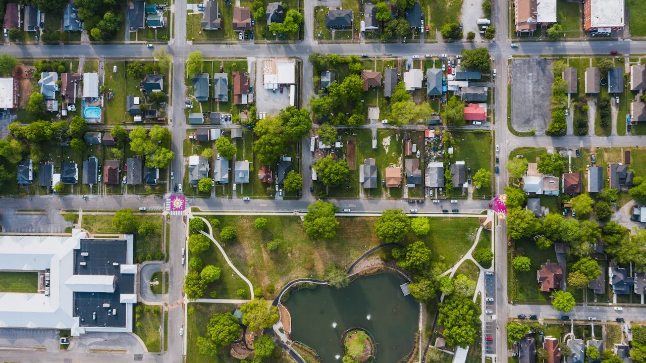 High-angle shot of a suburban neighborhood with streets, houses, and a park with a pond, showcasing vibrant greenery.